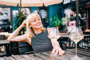 Happy smiling adult mature woman sitting in bar outdoors with wine glasses and blurry restaurant background scene, drinking white wine. Summer sunny day on patio. People lifestyle