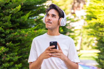 Young caucasian man in a park listening music with a mobile and thinking