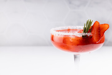Fresh classic strawberry margarita cocktail with green rosemary twig, ice cube, fruit slices, sugar rim in modern white kitchen interior on wooden table in sunlight, closeup.