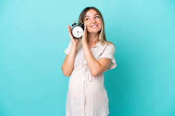 Fototapeta premium Young Romanian woman isolated on blue background pregnant and holding clock