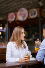 Charming smiling young woman enjoying fresh drink when sitting at cafe table with boyfriend