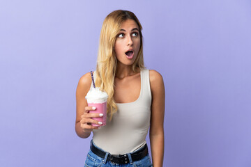Young blonde Uruguayan woman with strawberry milkshake isolated on purple background looking up and with surprised expression
