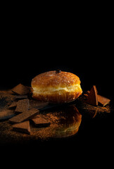 one large donut lies on a glass table with pieces of dark chocolate with reflection on a black background.