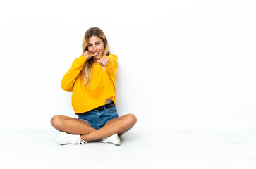 Young blonde Uruguayan woman sitting on the floor isolated on white background making phone gesture and pointing front