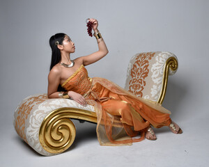 Full length portrait of pretty young asian woman wearing golden Arabian robes like a genie, seated pose on lounge, isolated on studio background.