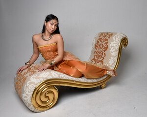 Full length portrait of pretty young asian woman wearing golden Arabian robes like a genie, seated...
