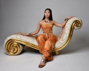 Full length portrait of pretty young asian woman wearing golden Arabian robes like a genie, seated pose on lounge, isolated on studio background.