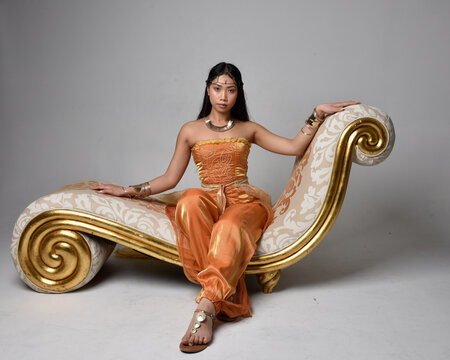 Full length portrait of pretty young asian woman wearing golden Arabian robes like a genie, seated pose on lounge, isolated on studio background.