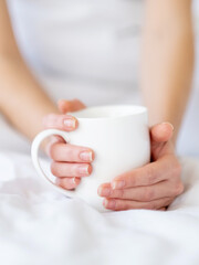 Fototapeta na wymiar Sunday coffee. Good morning. Home relaxation. Hot beverage. Closeup of elegant woman hands holding cup of latte drink on soft clean white bedsheets blanket in cozy bed.