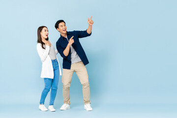 Full length portrait of happy excited Asian couple tourists pointing hands to empty space on isolated light blue background - Powered by Adobe