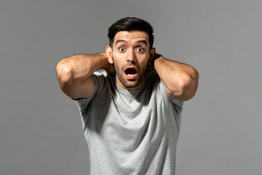 Shocked young Caucasian man gasping with hands around neck looking at camera on light gray studio background