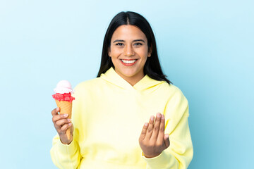 Young Colombian woman with a cornet ice cream isolated on blue background inviting to come with hand. Happy that you came