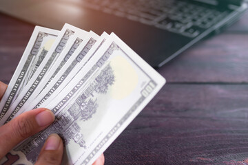 Finger holding bank note 100 dollars with keyboard laptop computer blur on wooden table. planing earn money investment and saving money future for 2022 new year. finance and business. count money pay