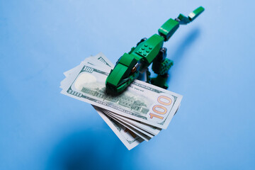 A pack of 100 dollar bills in mouth of tyrannosaur green kid toy isolated on blue background. Copy...