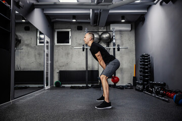 Fototapeta na wymiar Dead lifting workout with a kettle bell. Man in sportswear with a black T-shirt stands with his legs spread and lifts a kettle bell with both arms in gym. Strength and physical endurance