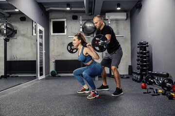 Fototapeta na wymiar Barbell squats with the help of personal training. Fit woman in sportswear and in good shape does barbell squats to strengthen the muscles of the whole body. Coach assistance in individual training