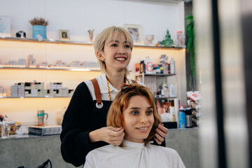 Portrait of happy young Asian woman hairstylist in apron while doing hairdressing and put care...