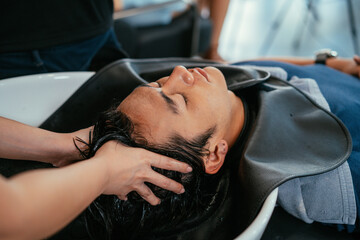 Handsome young Asian man relaxing with eyes closed with head in sink while hands of female hairdresser rinsing hair and pressing pressure points