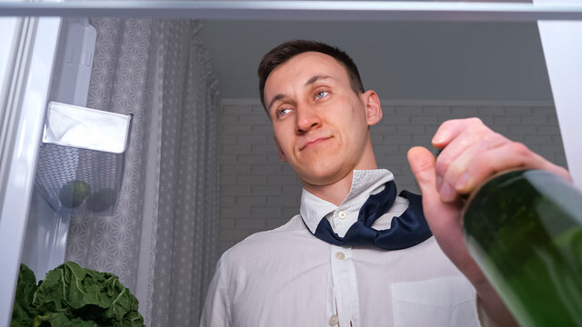 Drunk Man In White Shirt And Tie Opens Door To Takes Bottle Of Cold Beer From Shelf In Dark Kitchen, Close View From Inside New Refrigerator