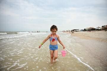 Pretty little girl holds a pink toy bucket in her hand and walks along the seacoast, enjoying sunbathing, outdoors recreation. Concepts of happy and carefree summer vacations at the beach