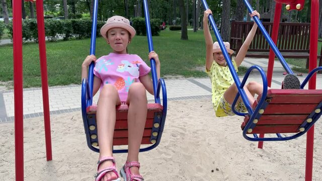 4k Cheerful twin sisters swinging on a swing on a playground.