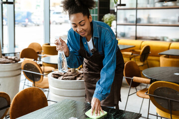 Black waitress wearing apron cleaning table while working in cafe