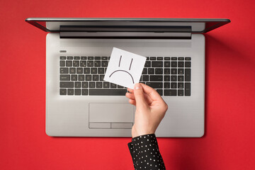 First person top view photo of hand holding white sticker note paper with drawn sad face over open...