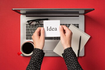 First person top view photo of female hands holding sticker note paper with inscription idea over open grey laptop glasses and red cup of coffee on isolated red background