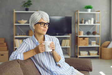 Portrait of pensive white haired lady drinking tea from cup in hands. Happy relaxed mature woman in glasses sitting on sofa at home, enjoying good cuppa coffee, looking away and thinking about future