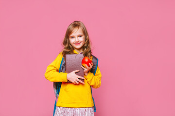 Back to school. Happy blonde girl  child  holds a book,  a red apple in her hand looking at the camera ​on a pink background. Education and intellectual development of children. World book day.