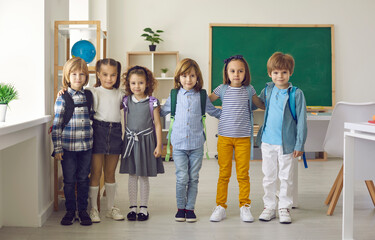 Portrait in the classroom of a small group of elementary school or kindergarten students. Kids in casual clothes and with backpacks on their shoulders are standing hugging and looking at the camera.