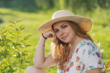 Cool young woman wearing straw hat is posing outdoors in summer day. Horizontally. 
