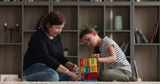 Happy loving older mature grandmother playing toys with preteen granddaughter, constructing building with plastic cubes, having fun entertaining together sitting on floor carpet in living room.