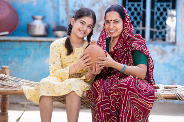 Portrait of happy rural indian mother and adorable daughter holding piggy bank while sitting on traditional bed at village home, Cute little girl collect money, saving and investment concept.