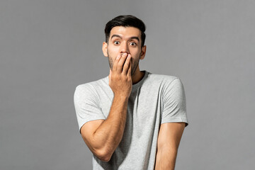 Portrait of shocked young Caucasian man with hand covering mouth on light gray studio background