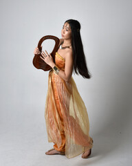 Full length portrait of pretty young asian woman wearing golden Arabian robes like a genie, holding...