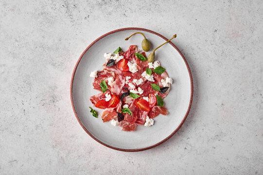 Prosciutto coppa di parma ham slices with ricotta cheese, tomato, capers and basil on light plate, top view, copy space