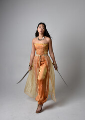 Full length portrait of pretty young asian woman wearing golden Arabian robes like a genie, holding a sword weapon, isolated on studio background.