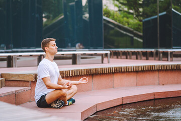 Attractive man athlete wearing sportswear meditate sitting in lotus pose in modern park at summer morning