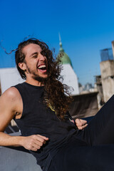 young brazilian man with long blond hair and curls laughing out loud on the terrace, sunbathing, city background. vertical photo