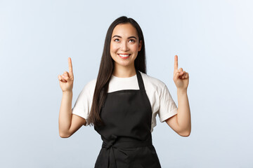 Coffee shop, small business and startup concept. Smiling cute asian female barista in black apron seeing something interesting on top banner. Girl working as cafe staff or waiter pointing fingers up