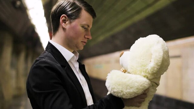 A young guy is talking with a toy bear at a transport stop. He's going on a date.
