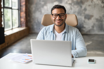 Cheerful indian male student or office worker is using laptop computer sitting at the desk. Friendly and optimistic eastern dark-haired freelancer guy in eyeglasses looks at camera and smiles