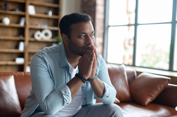 Full of doubts Indian guy sitting on the sofa at home, frustrated pensive eastern man worried about...