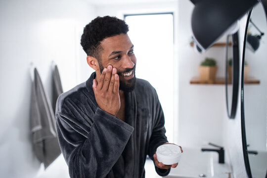 Young Man Applying Cream On Face Indoors At Home, Morning Or Evening Routine Concept.