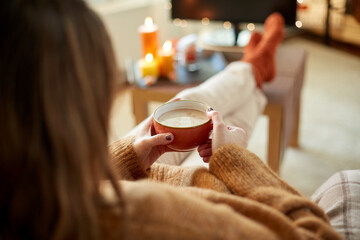 halloween, holidays and leisure concept - close up of young woman watching tv and drinking hot chocolate with her feet on table at cozy home