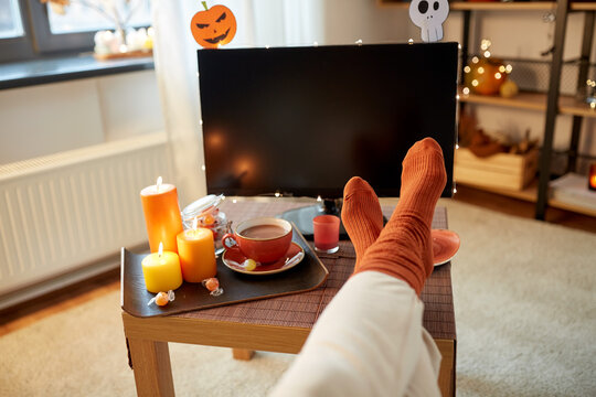 halloween, holidays and leisure concept - young woman watching tv and resting her feet on table at cozy home