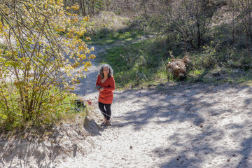 Naklejka premium Senior woman standing on a sandy path surrounded by vegetation, with her mobile phone gimbal tripod head stabilizer in hand, Zeepeduinen dunes in Burgh-Haamstede on a sunny day, Zeeland, Netherlands