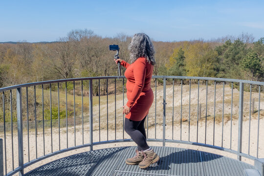 Woman filming with her mobile phone gimbal tripod head stabilizer the dunes of Zeepeduinen on metal viewpoint, with bare trees in the background on a sunny day in Burgh-Haamstede, Zeeland, Netherlands