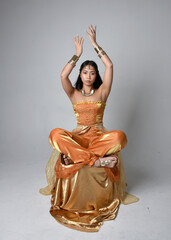 Full length portrait of pretty young asian woman wearing golden Arabian robes like a genie, seated pose, isolated on studio background.
